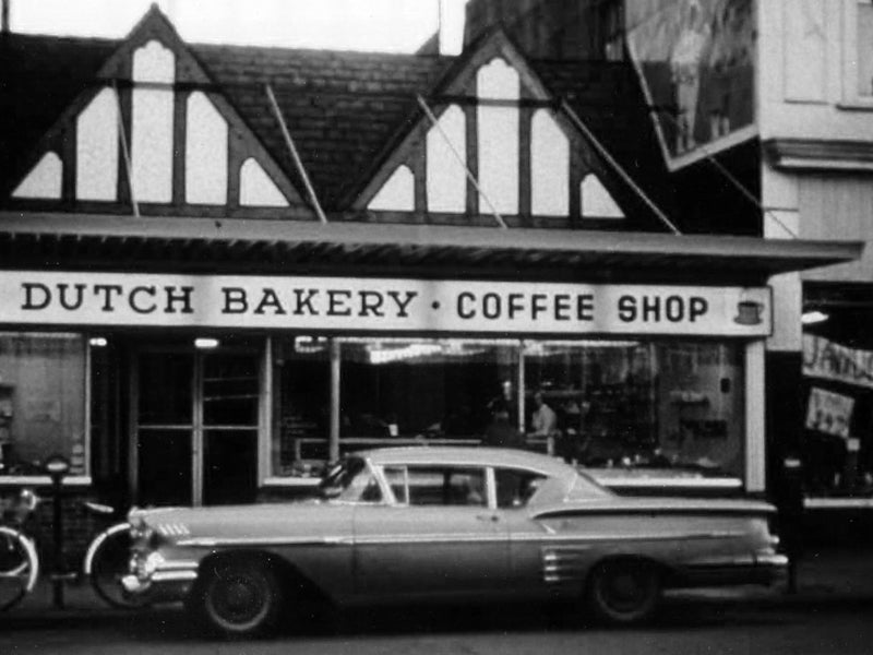 A black and white photo of the bakery store front with an updated sign and a vintage car parked in front.