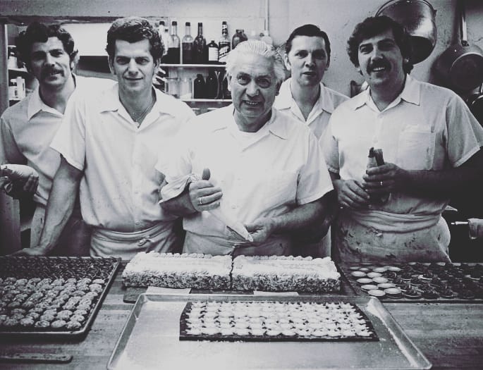 The original five men that started the Dutch Bakery standing behind a work top. Kees Sr is standing in the middle holding a piping bag while in the middle of decorating a cake